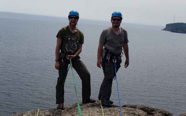 two people in rock climbing gear smile at the camera while standing on a cliff high above a body of water